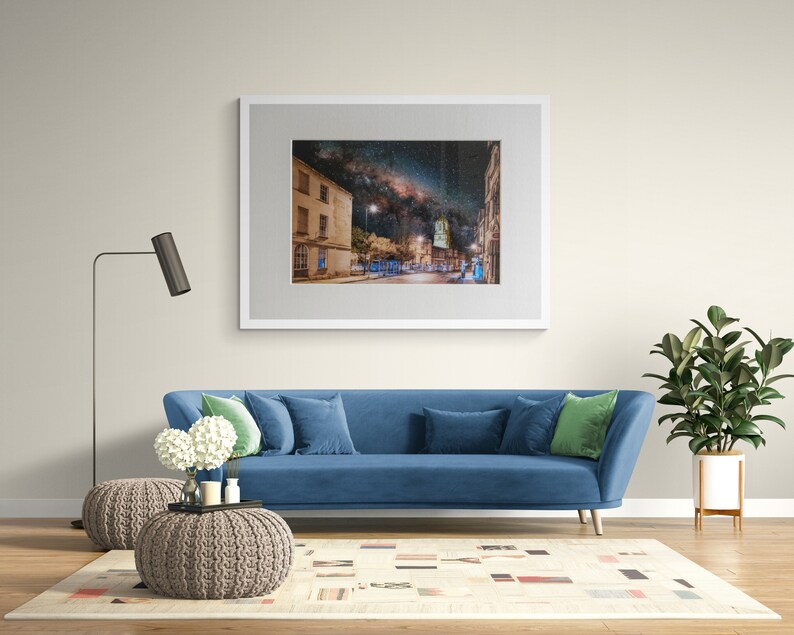 May include: A blue sofa with green throw pillows sits in a living room with a framed photograph of a city street at night with the Milky Way visible in the sky above. The sofa is in front of a white rug with a geometric pattern. A floor lamp with a black shade stands to the left of the sofa. A potted plant is on the right side of the sofa.