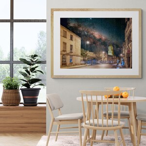 May include: A framed photograph of a city street at night with the Milky Way visible in the sky. The street is lined with buildings, and there are streetlights illuminating the scene. The photo is in a light wood frame.