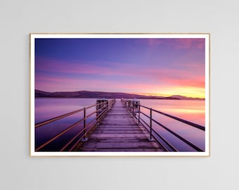 May include: A wooden pier extends out into a body of water at sunset. The sky is a vibrant mix of pink, purple, and orange. The water is calm and reflects the colors of the sky.