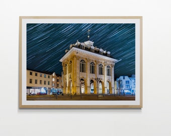 May include: A night time photo of a large, yellow building with a star trail in the sky above. The building has a clock tower and is lit up with lights. There are other buildings in the background.