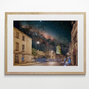 May include: A night time view of a street in a city with a tall building in the distance. The Milky Way is visible in the sky above the street. The street is lined with buildings and trees. The streetlights are on and there are cars parked on the side of the street.