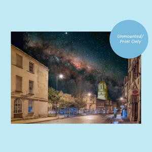 May include: A night time view of a street in a city with a tall building in the distance. The Milky Way is visible in the sky above the street. The street is lined with trees and streetlights.