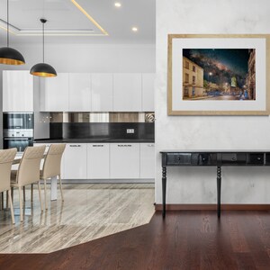 May include: A black console table with three drawers and four legs stands in front of a white wall with a framed print of a night sky over a city street. The table is in a modern kitchen with white cabinets and a marble floor.