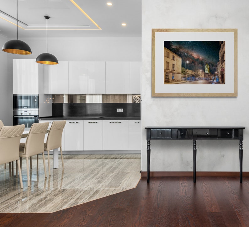 May include: A black console table with three drawers and four legs stands in front of a white wall with a framed print of a night sky over a city street. The table is in a modern kitchen with white cabinets and a marble floor.