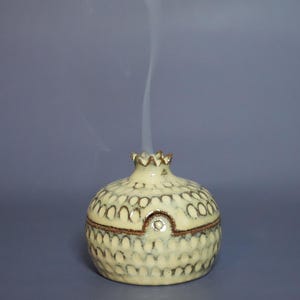 May include: A cream-colored ceramic incense burner shaped like a pomegranate with a textured, repeating scalloped pattern and a brown rim. A thin, vertical trail of white smoke rises from the top opening of the rounded, lidded vessel.