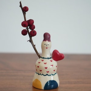 May include: A ceramic figurine of a woman with a red heart in her hand. She is wearing a white dress with red hearts and a red hat. The figurine is standing on a wooden surface.