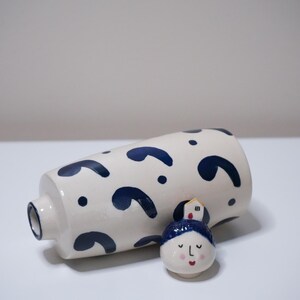 May include: A white ceramic vase with navy blue abstract shapes and a small, decorative face figurine with a blue hat and a house on top. The vase is cylindrical with a small opening.