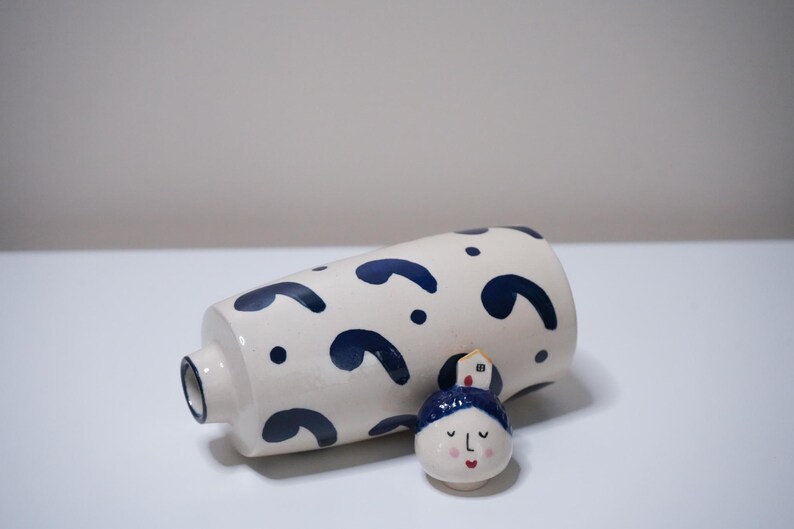 May include: A white ceramic vase with navy blue abstract shapes and a small, decorative face figurine with a blue hat and a house on top. The vase is cylindrical with a small opening.