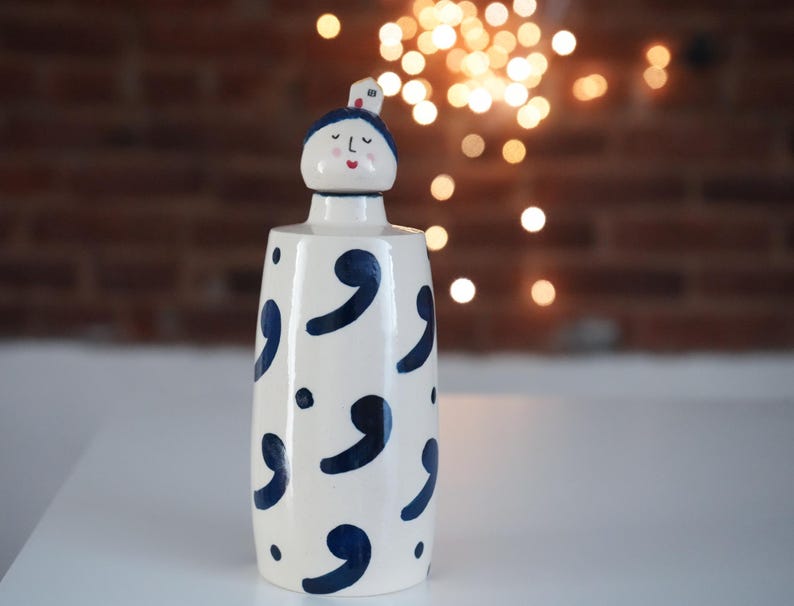May include: A white ceramic vase with a whimsical design. The vase features a blue comma pattern and a decorative head-shaped stopper with a painted face and a small house on top. The background includes a brick wall and bokeh lights.