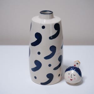 May include: A white ceramic vase with navy blue comma-shaped designs and dots. Beside it is a small, round ceramic figure with a face and a house on its head. The vase has a narrow neck.
