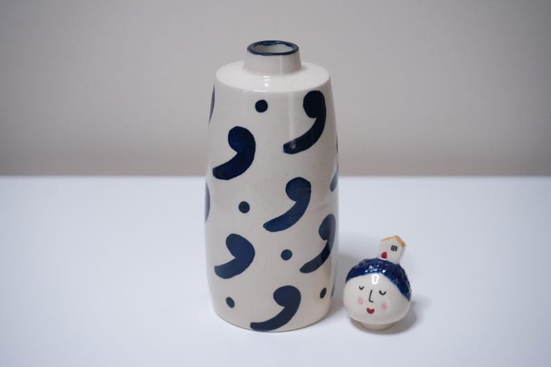 May include: A white ceramic vase with navy blue comma-shaped designs and dots. Beside it is a small, round ceramic figure with a face and a house on its head. The vase has a narrow neck.
