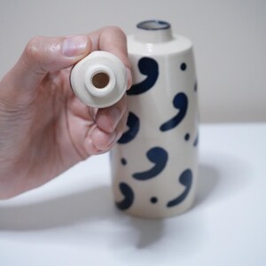 May include: A cream-colored ceramic vase with navy blue comma-shaped designs. The vase has a cylindrical shape and a small opening at the top. A matching stopper is held up to the vase.