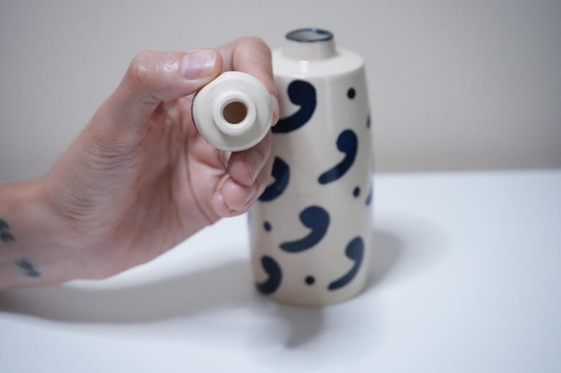 May include: A cream-colored ceramic vase with navy blue comma-shaped designs. The vase has a cylindrical shape and a small opening at the top. A matching stopper is held up to the vase.