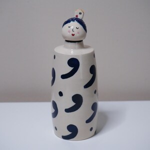 May include: A ceramic decorative bottle with a whimsical design. The bottle is off-white with navy blue comma-shaped patterns. The top features a stylized face with blue hair and a small house. The bottle is on a white surface.