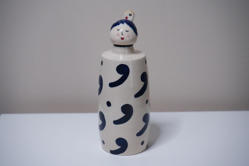May include: A ceramic decorative bottle with a whimsical design. The bottle is off-white with navy blue comma-shaped patterns. The top features a stylized face with blue hair and a small house. The bottle is on a white surface.