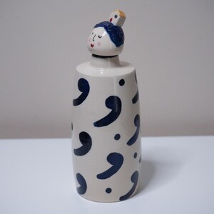 May include: A ceramic bottle with a decorative design. The bottle is off-white with navy blue comma shapes and dots. The top features a small face with blue hair and a small square on top.