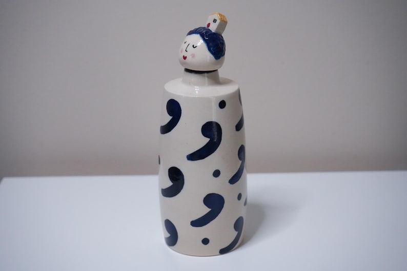 May include: A ceramic bottle with a decorative design. The bottle is off-white with navy blue comma shapes and dots. The top features a small face with blue hair and a small square on top.