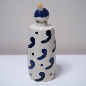 May include: A white ceramic bottle with a dark blue comma pattern and a dark blue and white stopper. The bottle has a cylindrical shape and is suitable for storing liquids. The stopper is made of ceramic and cork.