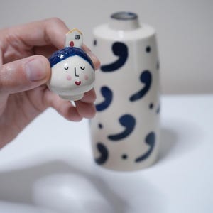 May include: A small, hand-painted ceramic figurine with a face and a house on top is held near a matching vase. The figurine has a blue head covering, closed eyes, and a red mouth. The vase is off-white with navy blue comma-shaped designs.