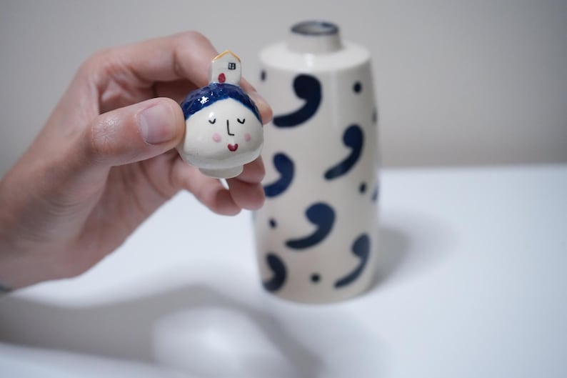 May include: A small, hand-painted ceramic figurine with a face and a house on top is held near a matching vase. The figurine has a blue head covering, closed eyes, and a red mouth. The vase is off-white with navy blue comma-shaped designs.