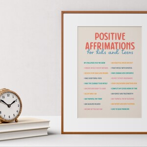 May include: A framed print with the text "POSITIVE AFFIRMATIONS for kids and teens" in red and blue. A gold-colored woven clock sits on a stack of books to the left of the print.
