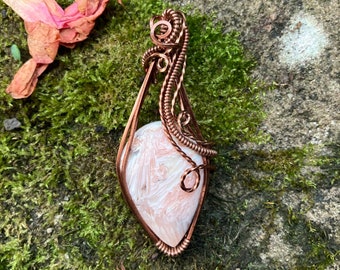 May include: A handcrafted copper wire pendant featuring a teardrop-shaped stone with a white and pink pattern. The wire is intricately woven around the stone, creating a decorative frame. The pendant is displayed on a bed of green moss.