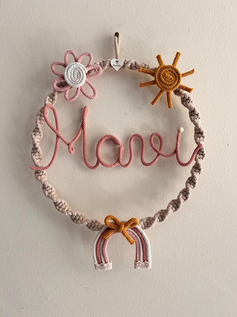 May include: A handmade macrame wreath with the name "Marie" in pink yarn. The wreath features a flower, sun, and rainbow, all in shades of pink, white, and yellow. A small wooden heart is attached at the top.