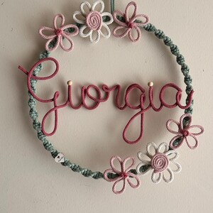 May include: A handmade macrame wreath with the name "Giorgia" in pink script. The wreath is adorned with pink and white flowers and a green rope border. A decorative piece for a nursery or bedroom.