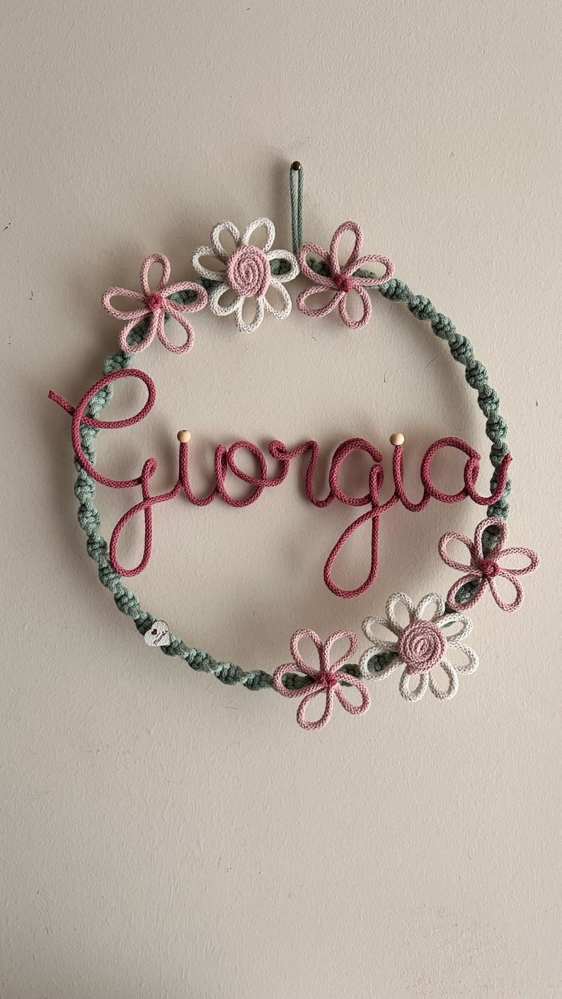 May include: A handmade macrame wreath with the name "Giorgia" in pink script. The wreath is adorned with pink and white flowers and a green rope border. A decorative piece for a nursery or bedroom.