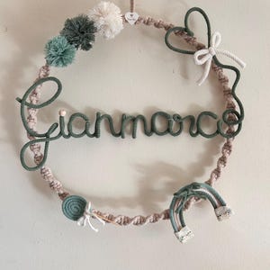Può includere: Una ghirlanda in macramè fatta a mano con il nome "Gianmarco" in filo verde. La ghirlanda presenta una varietà di elementi decorativi, tra cui pompon nei toni del verde e del bianco, un piccolo arcobaleno e un accento a forma di cuore.