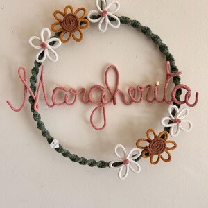 May include: A handmade wreath with the name "Margherita" in pink yarn. The wreath is adorned with crocheted flowers in white, brown, and yellow, set against a green rope base. A charming, decorative piece for home decor.