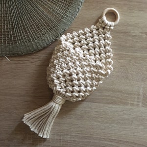 Può includere: Un arazzo da parete in macramè bianco con un anello di legno in alto e un lungo fiocco in basso.