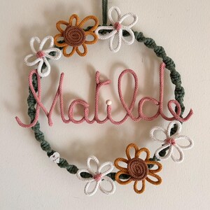 May include: A handmade wreath with the name "Matilde" in pink yarn. The wreath is decorated with white, brown, and yellow crocheted flowers and a green yarn base. A loop of green yarn is at the top for hanging.
