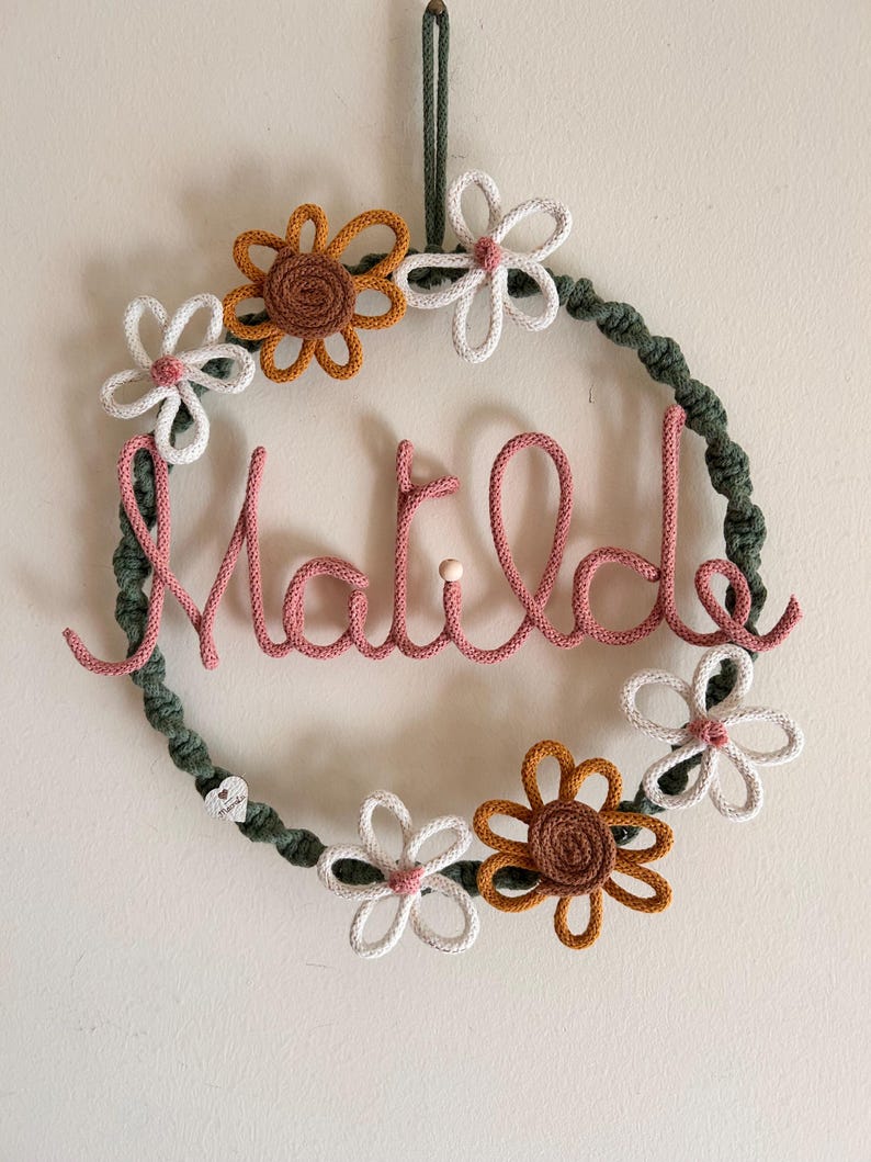 May include: A handmade wreath with the name "Matilde" in pink yarn. The wreath is decorated with white, brown, and yellow crocheted flowers and a green yarn base. A loop of green yarn is at the top for hanging.