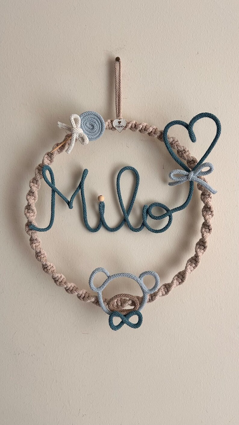 May include: A handmade macrame wall hanging in a circular shape, featuring the name "Milo" in teal blue. The wreath includes a bear, heart, and flower accents, all in shades of blue and beige. A wooden bead is also included.