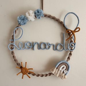 May include: A handmade macrame wall hanging in a circular shape, featuring the name "Leonardo" in light blue script. The wreath is adorned with blue and white pom-poms, a sun, a rainbow, and a bow. The overall color scheme is neutral with blue accents.