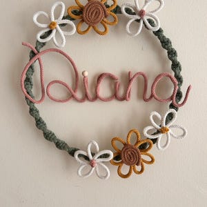 Può includere: Una ghirlanda in macramè fatta a mano con il nome "Diana" in filo rosa. La ghirlanda è decorata con fiori in filo bianco, giallo e marrone. È sospesa da un cordoncino verde.
