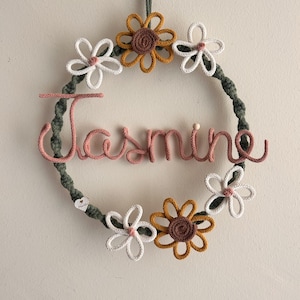 May include: A handmade floral wreath with the name "Jasmine" in pink script. The wreath features a green rope base adorned with white, yellow, and brown crocheted flowers. A decorative piece for a nursery or bedroom.