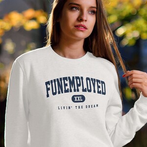May include: White crew neck sweatshirt with the text "FUNEMPLOYED" in navy blue, size XXL. The text "Livin' the Dream" is below the main text. The person is wearing blue jeans and white sneakers.