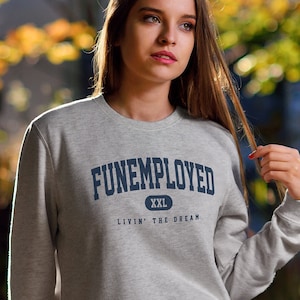 May include: A light gray sweatshirt with the text "FUNEMPLOYED" in navy blue, with "XXL" below it and "LIVIN' THE DREAM" underneath. The person is wearing blue jeans and white sneakers.