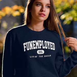 May include: Navy blue sweatshirt with the text "FUNEMPLOYED" in white block letters. Below the main text is "XXL" and "LIVIN' THE DREAM". The sweatshirt is paired with blue jeans.