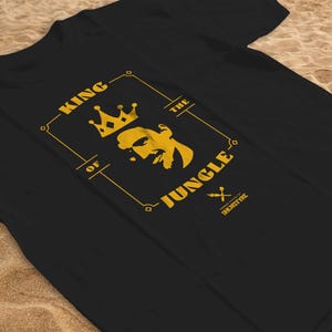 May include: A black t-shirt featuring a gold graphic print of a lion's head wearing a crown, enclosed in a rectangular frame. The text reads KING OF THE JUNGLE, with smaller text below that says SHURITYRE. The design is centred on the chest area.