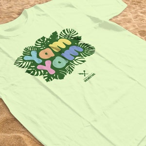 Puede incluir: Una camiseta verde claro con un gráfico de la palabra "yam" en letras de colores rodeada de hojas verdes. El texto "THE SWEET SPOT" está impreso en letras pequeñas debajo del gráfico.