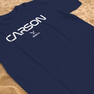 Puede incluir: Una camiseta azul marino con la palabra "CARSON" impresa en blanco en la parte delantera. El texto está en negrita, fuente sans-serif. Debajo de la palabra "CARSON" hay un pequeño logotipo blanco con una cruz y el texto "SECURITY INC."