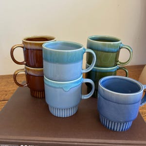 May include: A collection of six ceramic mugs with a two-tone glazed finish and a ribbed base. The mugs are arranged in stacks and feature various colours including brown, light blue, and green. Each mug has a handle and a smooth, glossy rim.