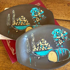 May include: Two oval-shaped brown serving trays with a design of a wheelbarrow filled with yellow and brown squares, a bunch of grapes, and wheat stalks. The wheelbarrow is painted turquoise.