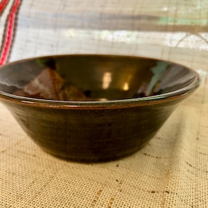 May include: A dark brown ceramic bowl with a slightly raised rim. The bowl has a subtle sheen and is sitting on a beige textured surface.