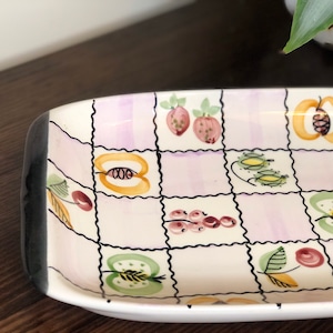 May include: A white ceramic rectangular serving dish with a black rim. The dish has a checkered pattern in shades of pink and white with a variety of fruit designs in yellow, green, red, and black.