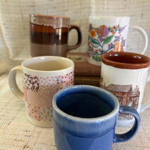 May include: Four ceramic mugs with different designs. One mug is brown with white stripes, one is white with floral designs, one is white with a pink and blue patchwork design, and one is blue with a ribbed design.
