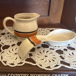 May include: A cream-coloured ceramic creamer jug and a matching spoon rest, both featuring a horizontal band design in dark brown and burnt orange. The jug has a curved handle and a spout, while the spoon rest has a long, curved handle.
